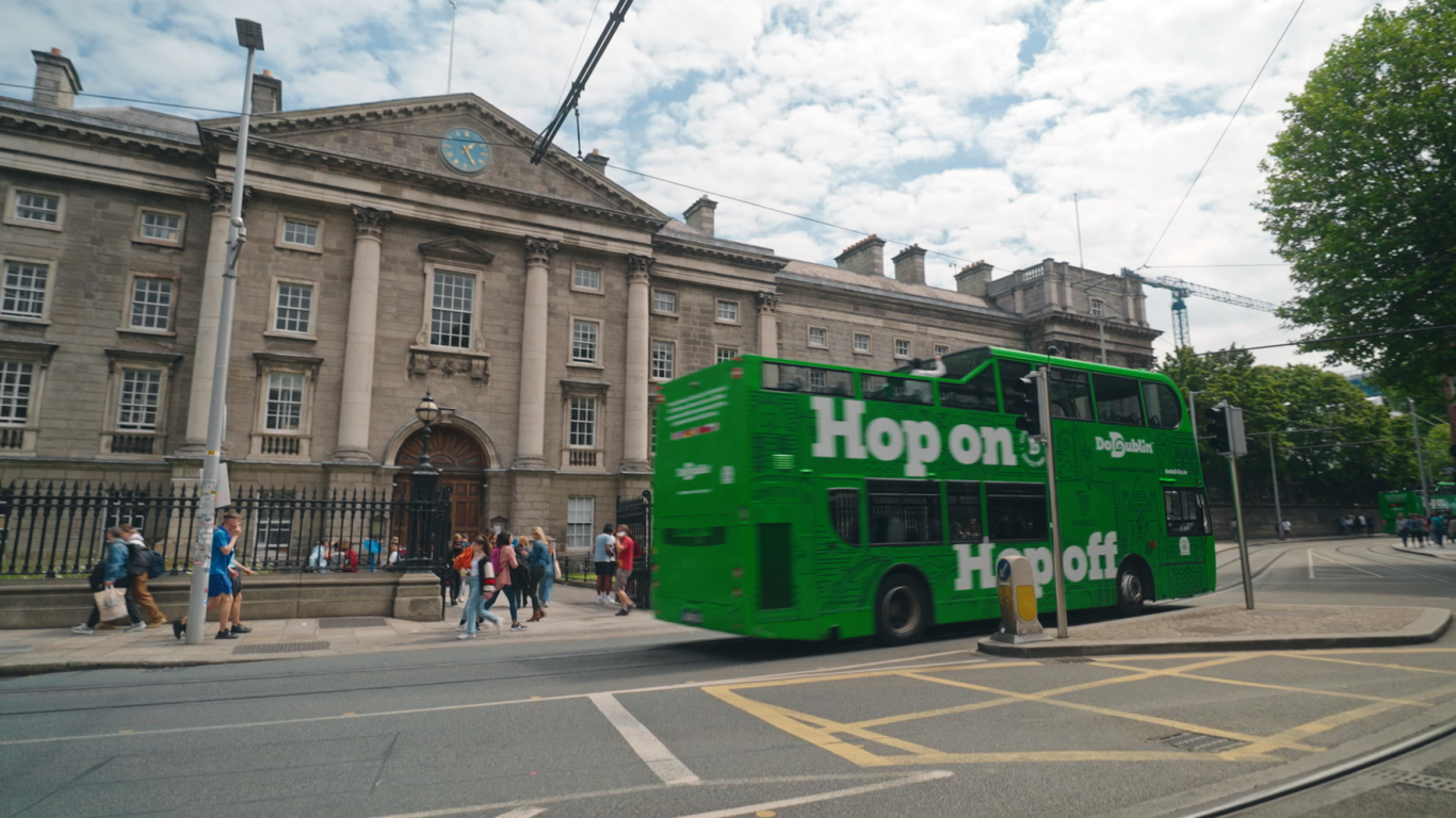 the tour bus on the road passing a building hop on hop off bus passing big building, can see back and side of the bus